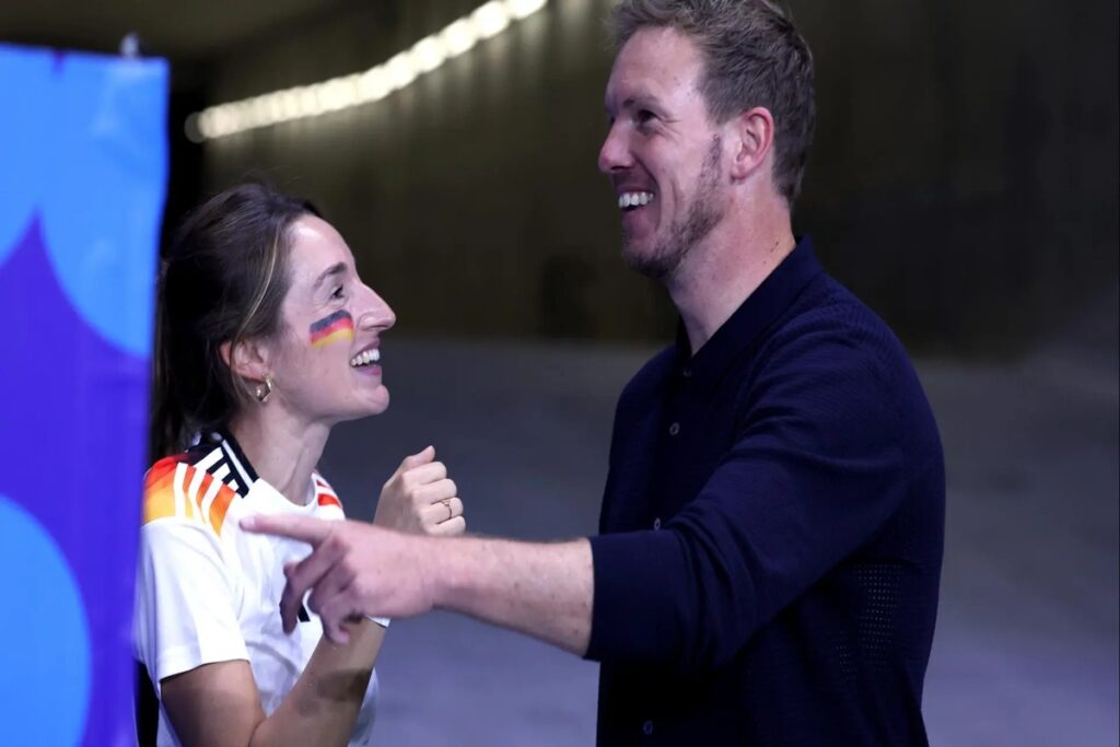 Lena Wurzenberger und Julian Nagelsmann lächelnd und vertraut bei einem EM-Spiel, Deutschland-Flagge auf ihrer Wange.